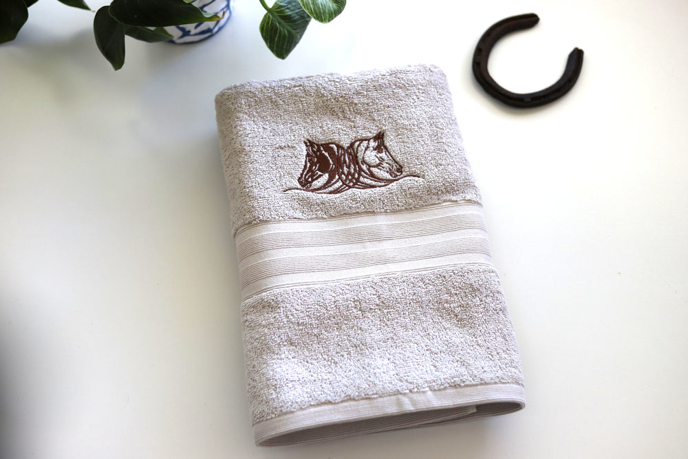 A fawn colored bath towel with two embroidered horse heads in chocolate colored thread