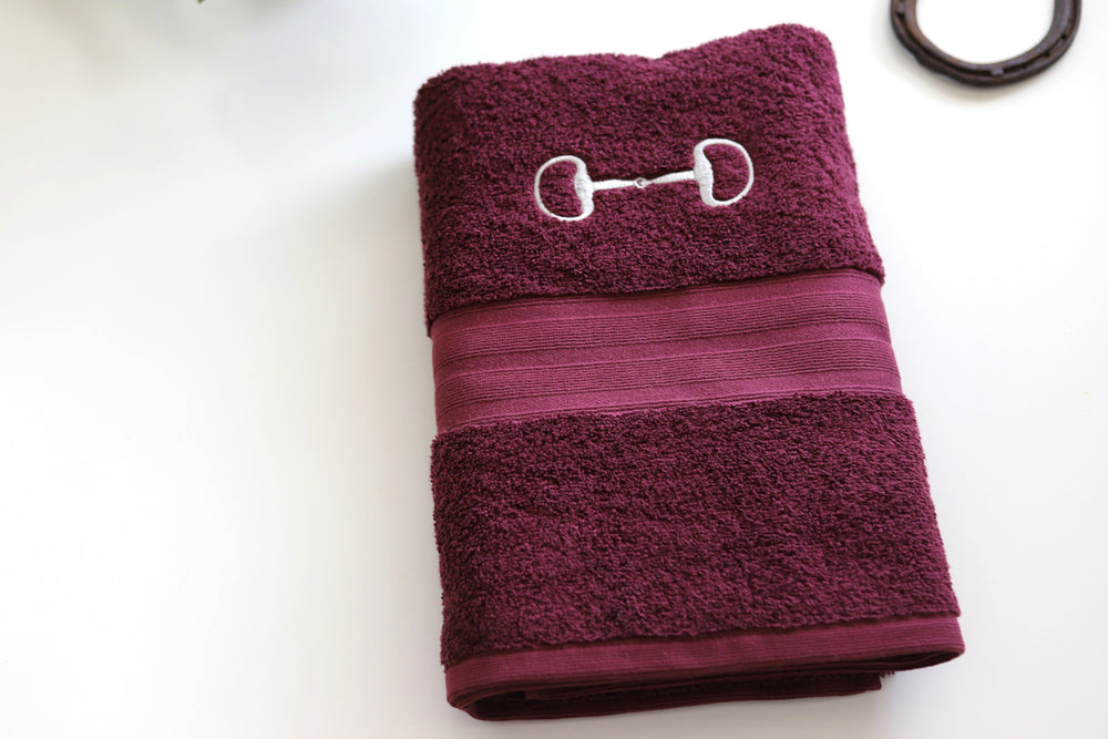 A burgundy bath towel with an embroidered snaffle bit design on the front.