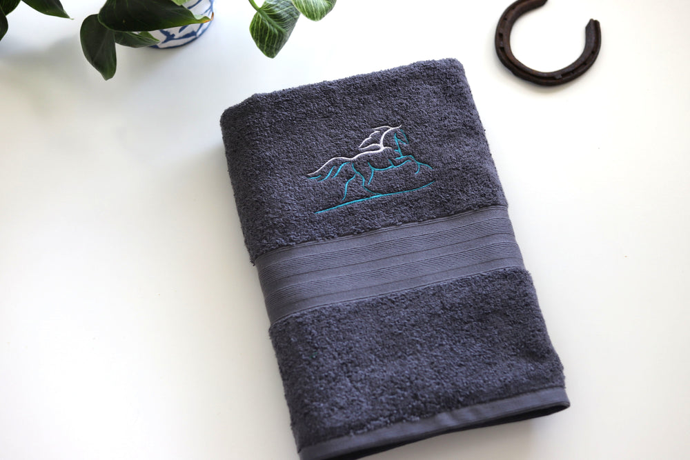 A steel grey combed cotton bath towel with an embroidered stylised horse outline design in teal and white thread