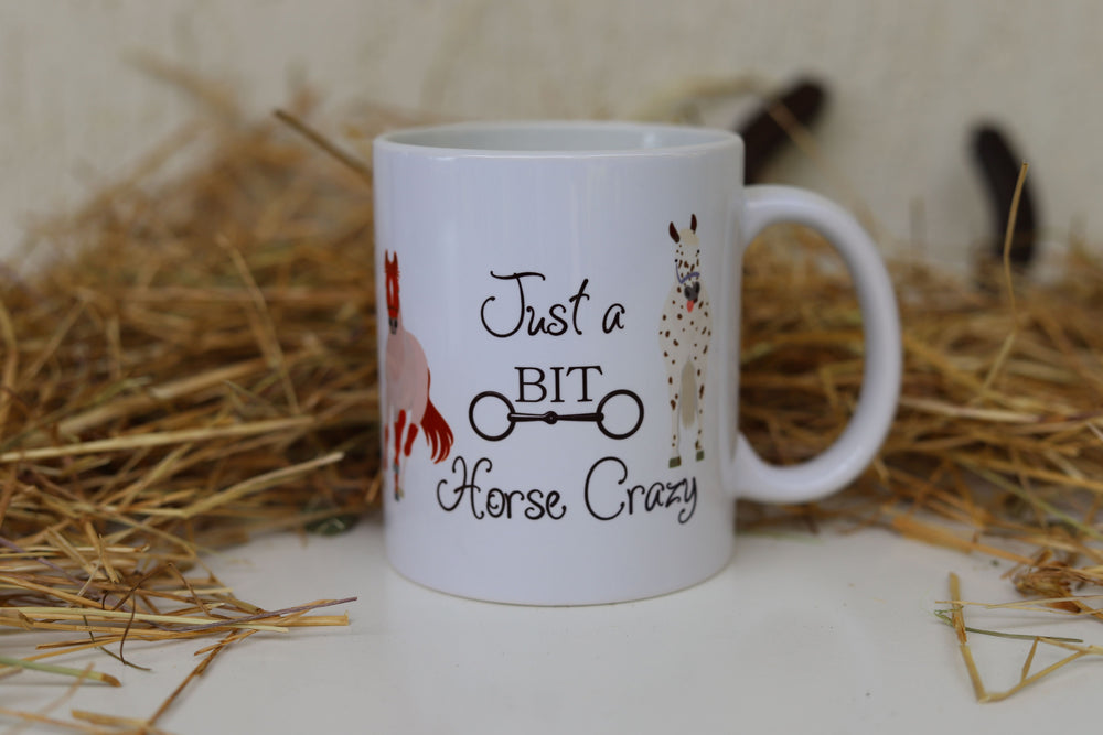 A white ceramic mug with 'Just a BIT Horse Crazy' text and horse illustrations placed on a straw background.