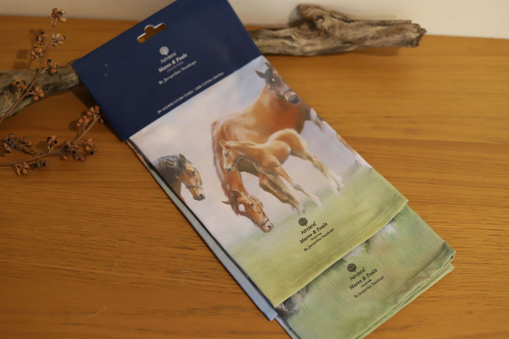 A set of two folded tea towels featuring an artistic print of horses, specifically mares and foals, in a pasture on a blue packaging card.