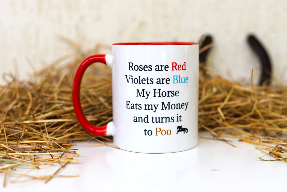 A white ceramic mug with red trim with text that reads 'ROSES ARE RED, VIOLETS ARE BLUE, MY HORSE EATS MY MONEY AND TURNS IT TO POO', placed on a straw bed 
