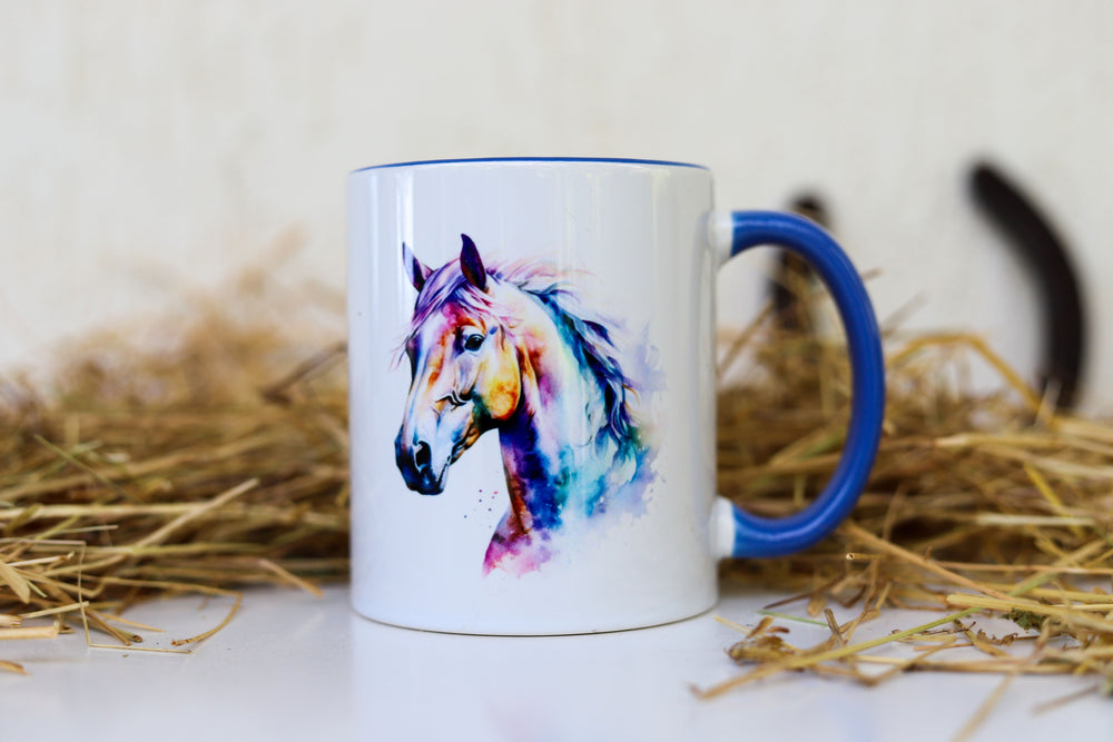 A ceramic mug with a watercolor print of a horse head on both sides, featuring a blue handle and placed on a surface with straw hay around it.