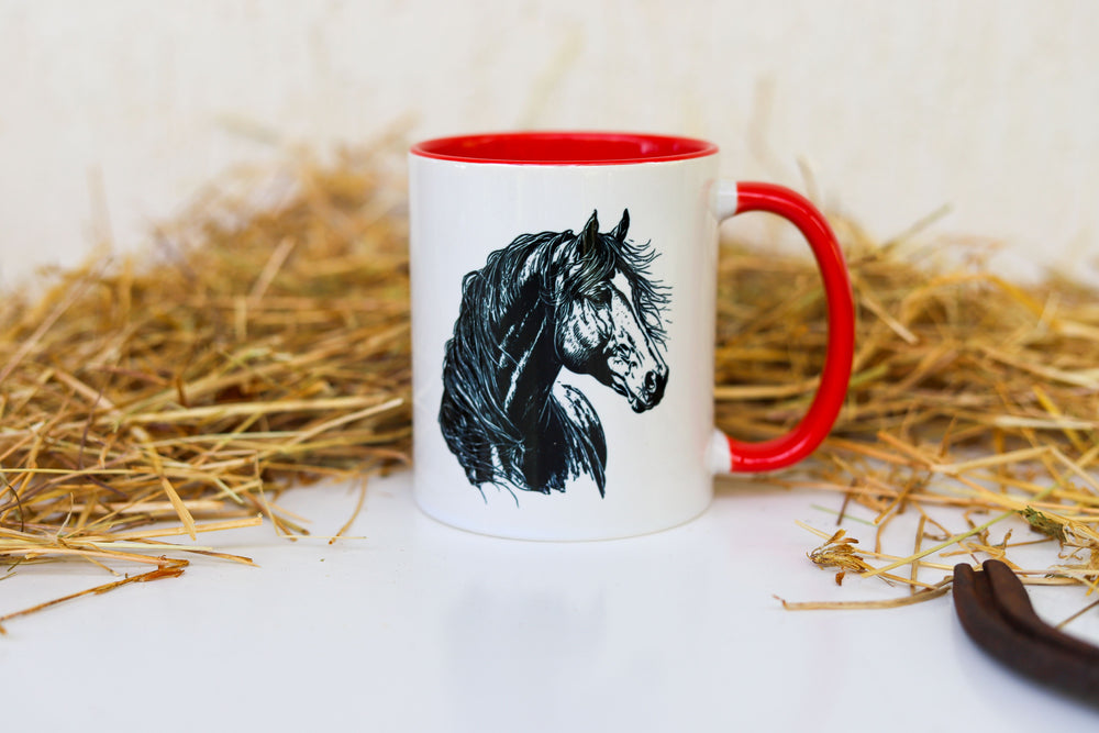 A white ceramic mug with red trim with a black horse head design, placed on a straw background.