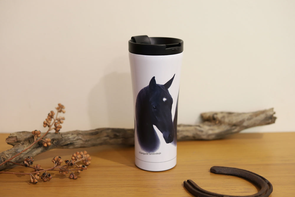 A stainless steel travel mug with a black horse design, featuring a double walled design, placed on a wooden surface with a branch and horseshoe nearby.
