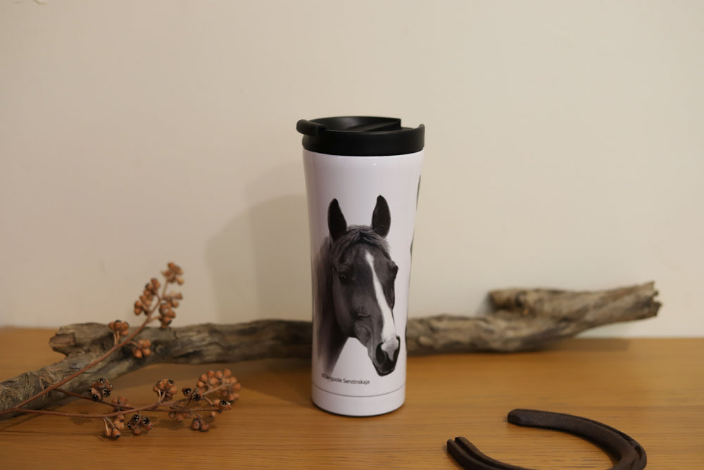 A travel mug with a chestnut horse design, placed on a wooden surface surrounded by props including a horseshoe.