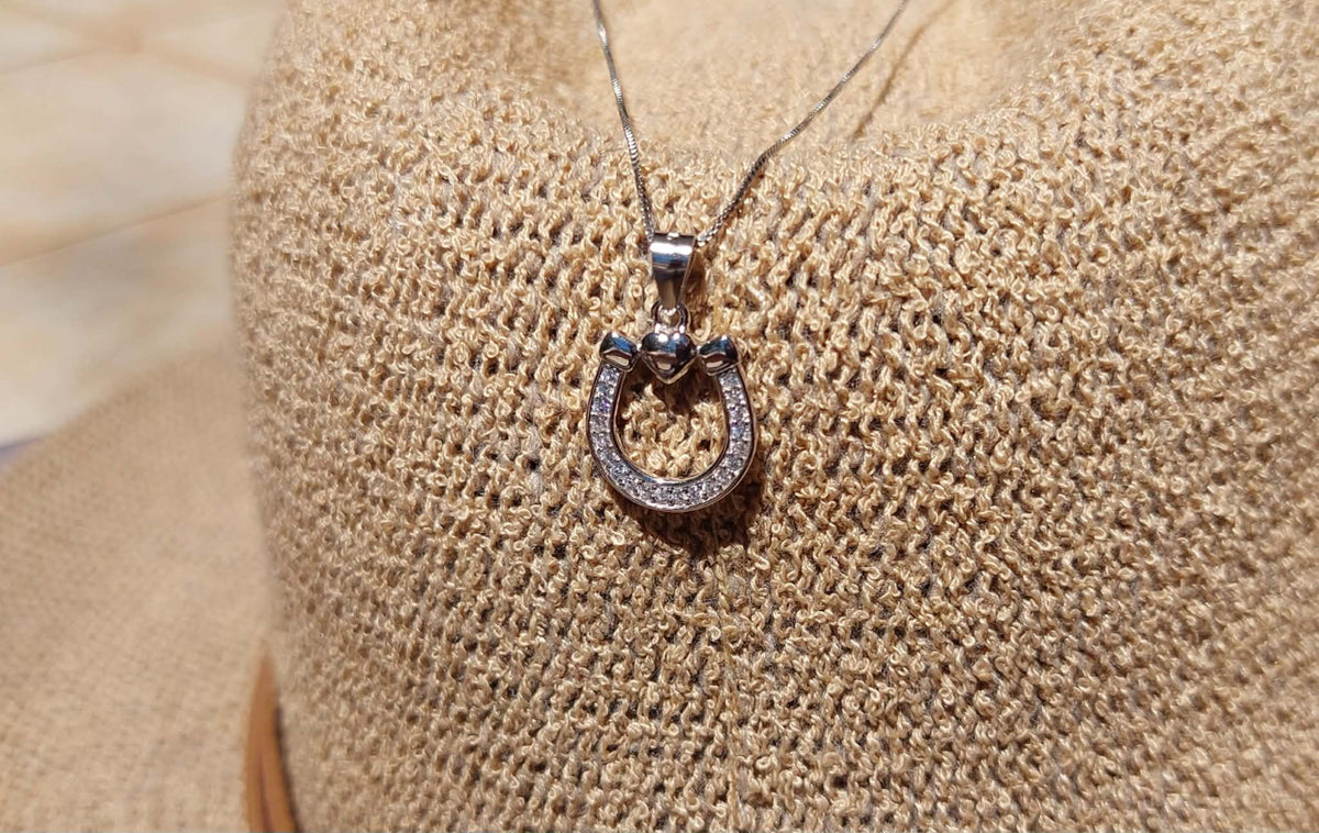 Sterling Silver Horseshoe and heart pendant on a sterling silver chain leaning on a woven beige hat as background 